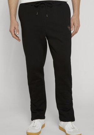 Uomo che indossa pantaloni della tuta neri con coulisse e scarpe da ginnastica bianche in piedi su sfondo semplice.