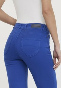Jean en denim bleu avec une coupe ajustée. Ses caractéristiques incluent des détails de couture discrets, deux poches arrière et une étiquette sur la taille.