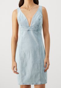 Vaaleansiniinen farkkupuku, jossa on syvä V-aukkoinen kaula-aukko, hihaton muotoilu, tiukka siluetti ja pystysuorat ompeluolkohdat.