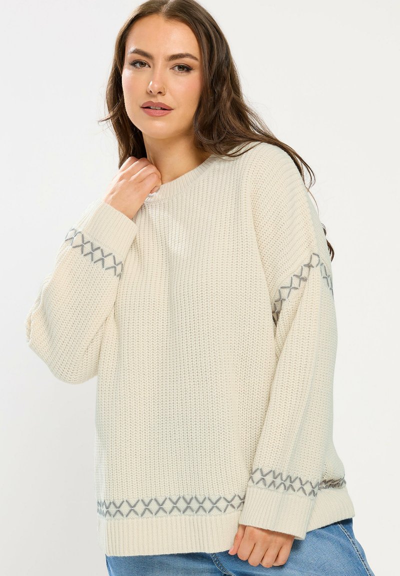 Giovane donna che indossa un maglione lavorato a maglia color crema con motivi incrociati grigi su maniche e bordo, abbinato a jeans blu, in piedi su sfondo bianco.