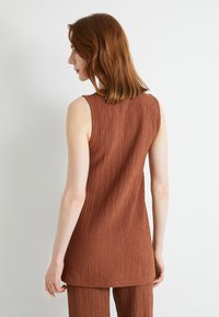 Bruine, mouwloze top van een gestructureerde stof. Het ontwerp heeft een ronde halslijn en een rechte zoom die net voorbij de heupen valt.