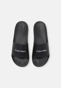 Sandali neri con la suola piatta realizzati in un materiale liscio, caratterizzati da una cintura larga con la scritta "Calvin Klein" in bianco, design minimalista.