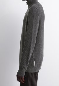 Pull en gris à côtes avec un col haut, des manches longues et une étiquette sur le côté. Présente un motif de tricot texturé et une coupe ajustée.