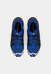 Kék sportcipők, melyek texturált szövet felsőrésszel, kontrasztos fekete részletekkel, valamint vastag, fehér és fekete talppal rendelkeznek, melynek tapadós profilja van.