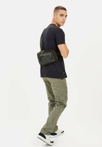Sort nylon skuldertaske med to lynlåslommer, justerbar rem og minimalistisk design; båret på ryggen.