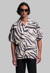 Camicia a maniche corte color crema con audaci strisce zebra nere, dotata di tasca sul petto e dal taglio morbido. Il tessuto sembra leggero e liscio.