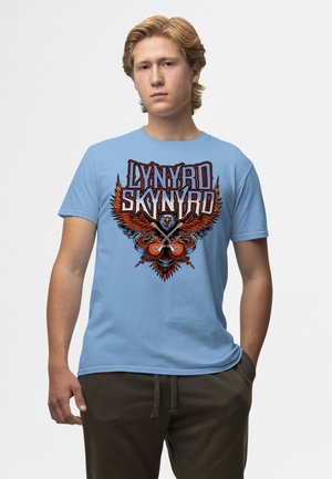 Junger Mann trägt ein hellblaues Lynyrd Skynyrd T-Shirt mit Adler und gekreuzten Gitarren Grafik, steht vor einem schlichten Hintergrund.