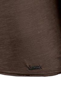 Braune Baumwollbluse mit glatter Textur, abgerundetem Saum und einem kleinen metallischen Markentag nahe dem unteren Rand.