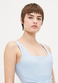 Maiô azul claro com decote quadrado e alças largas, feito de tecido liso. Apresenta um detalhe com nó na cintura.