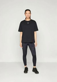 Sieviete stāv pretī vienkāršai fona, ģērbusies brīvā melnā T-kreklā, pieguļošos melnos legingos un melnās sporta apavās.