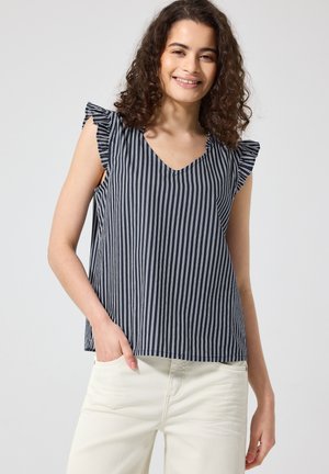 Joven mujer con pelo rizado que lleva una camiseta sin mangas de rayas verticales en blanco y negro y pantalones de color claro, sonriendo con una mano en el bolsillo.
