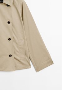 Manteau beige avec un col cranté, cinq boutons foncés et une poche latérale. Fabriqué en tissu lisse, avec une coupe légèrement ample.
