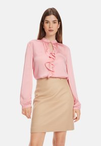 Helle rosa Bluse mit Rüschendetail an der Vorderseite, Schlüssellochöffnung und langen Ärmeln. Kombiniert mit einem beigen, taillierten Rock. Glatte Stoffoberfläche.