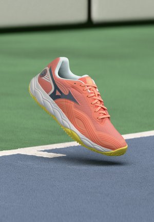 Zapatilla de correr coral y amarilla con logo negro flotando sobre una pista deportiva azul y verde con líneas blancas de delimitación.