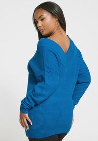 Niebieski sweter robiony na drutach z dekoltem w kształcie litery V, długimi rękawami i prążkowaną strukturą. Tył swetra ma wyraźne wycięcie w kształcie litery V dla dodatkowego detalu.