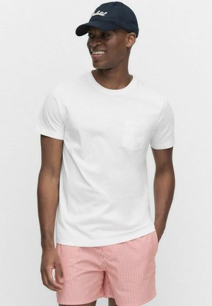 Hvid kortærmet t-shirt med en brystlomme, parret med røde og hvide stribede shorts. Fladskygget marineblå kasket. Enkel, afslappet design.