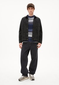 Schwarze Jeansjacke mit Druckknöpfen, über einem mehrfarbigen Strickpullover mit blauen und weißen Streifen, kombiniert mit lockeren dunklen Jeans und grauen Sneakern.