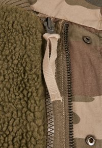 Zelená fleece tkanina vedľa béžovej camouflage vonkajšej časti, obsahujúca zips a tkaninový ťahák, s kovovými prvkami a detailom na zacvakávanie.
