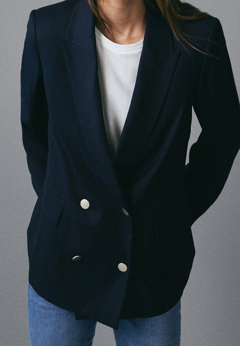 Persona che indossa un blazer doppiopetto blu navy con bottoni argentati sopra una camicia bianca e jeans blu, in piedi contro uno sfondo grigio.