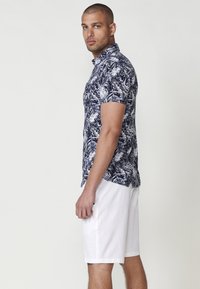 Majica s kratkimi rokavi v mornarsko modri barvi z belim vzorcem tropskih listov, v kombinaciji z belimi kratkimi hlačami. Majica ima ovratnik in zapenjanje na gumbe.