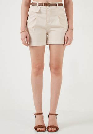 Vrouw draagt beige geplooide shorts met hoge taille, met een bruine riem en bruine gevlochten sandalen met enkelbandje, staand tegen een effen achtergrond.