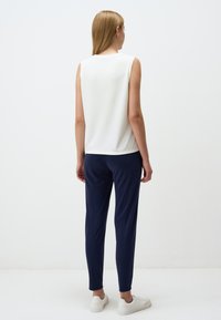 Witte mouwloze top gecombineerd met marineblauwe tapered broek. De broek heeft een gladde textuur en beschikt over een elastische tailleband. Model draagt witte sneakers.