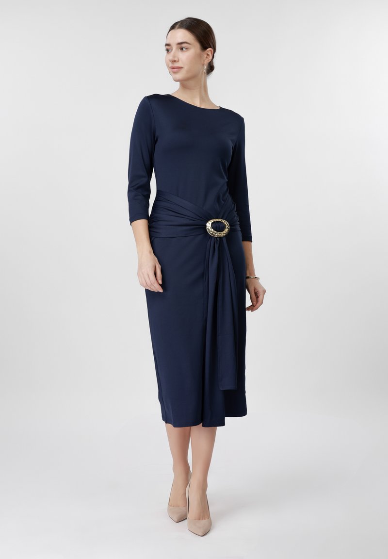 Woman in navy blue midi dress with three-quarter sleeves and a large decorative buckle on gathered fabric at the waist, wearing beige heels.