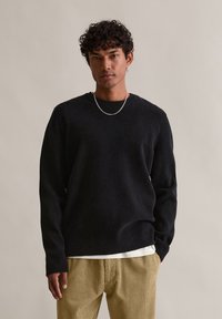 Μαύρος υφασμάτινος γυαλιστερός πουλόβερ με μακριά μανίκια, στρογγυλή λαιμόκοψη και ασημένια αλυσίδα ως αξεσουάρ, σε συνδυασμό με ριγωτό μπεζ παντελόνι.