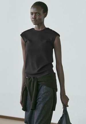 Donna con capelli corti e ricci, indossa una maglia nera senza maniche e pantaloni scuri, tiene in mano una borsa nera e ha una felpa scura annodata in vita.