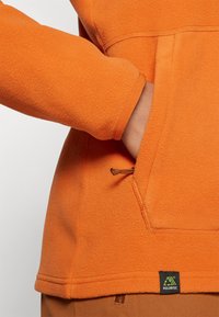 Pomarańczowy polarowy sweter z kieszenią typu kangur, z brązowym sznurkiem, szytymi szwami oraz metką Polartec u dołu.