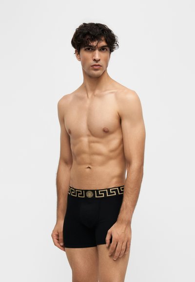 Jeune homme aux cheveux bouclés portant un boxer noir avec une taille à motif doré, debout devant un fond blanc uni.
