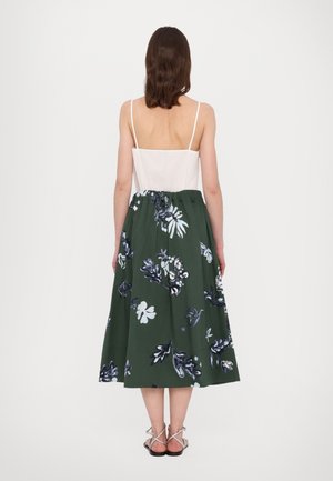 Femme portant un haut blanc à fines bretelles, une jupe midi verte avec des motifs floraux blancs et bleus marins, et des sandales plates argentées à brides, debout.