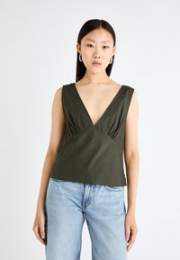 Olive green sleeveless top with a deep V-neck, horizontal gathers at the bust, and a loose fit, paired with light blue jeans.