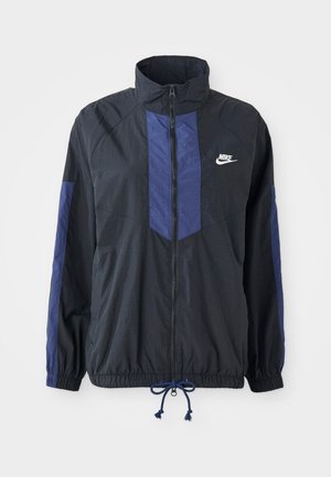 Svart jakke med marineblå detaljer, høy krage, full glidelås foran, elastiske mansjetter og snøring i nederkant. Nike-logo på venstre bryst. Lettstoff.
