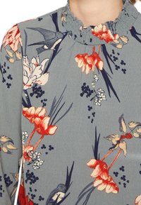 Blommig blus i mjuk blå tyg, med röda och krämfärgade blommor, gröna blad och en volangkrage. Livlig design med detaljerade mönster.