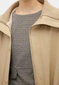 Chaqueta beige con cremallera y cuello alto combinada con un vestido estampado que presenta un diseño a cuadros en tonos marrones y negros. Se puede observar un notable detalle de botones.