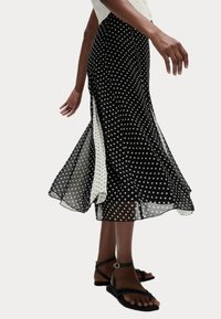 Jupe à pois noir et blanc avec un tissu transparent, une coupe évasée, des fentes latérales et une silhouette fluide. Portée avec des sandales à lanières noires.