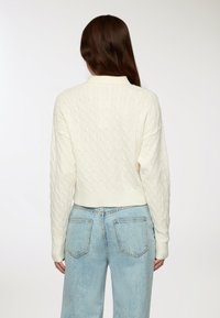 Donna che indossa un maglione bianco a trama a trecce e jeans di denim blu chiaro, vista da dietro, su uno sfondo neutro.