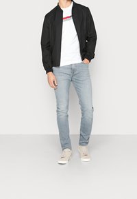 Chaqueta bomber negra sobre una camiseta blanca con rayas rojas y negras, combinada con jeans de color azul claro y zapatillas beige.
