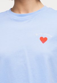 Ανοιχτό μπλε πουκάμισο με ένα μικρό κεντημένο κόκκινο καρδιά και λευκό κείμενο που λέει "Να είσαι ο Βαλεντίνος μου" στο επάνω μέρος του στήθους.