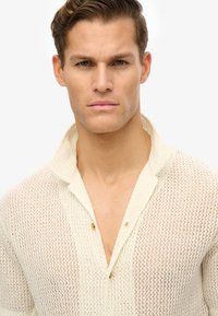 Strikskjorte i creme med en let, åndbar tekstur. Har en krave og tre knapper ved halsudskæringen, som viser et afslappet design.