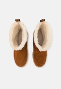 Stivali in suede marrone con un morbido rivestimento in crema, forma arrotondata e suola piatta in gomma, dotati di un pull tab per indossarli facilmente.