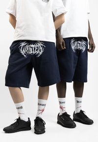 Shorts en coton bleu marine avec le logo "WASTE" en blanc, associés à des chaussettes blanches avec des accents rouges et des baskets noires. Design simple et décontracté.