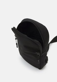 Svart stoffveske med glidelås på toppen, med en frontlomme og justerbar stropp, designet for å bære små gjenstander sikkert.