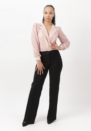 Frau steht vor weißem Hintergrund in rosa Bluse mit langen Ärmeln und aufgebauschten Schultern sowie schwarzer, hoch taillierter Hose.