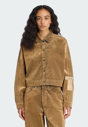 Chaqueta de pana marrón recortada con cierre de botones, costuras contrastantes y rayas laterales. Cuenta con un parche del logo de Adidas en la manga.