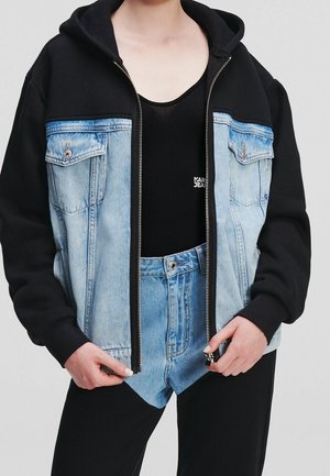 Personne portant une veste à capuche noire avec des panneaux avant en denim, un haut noir et un short en denim taille haute par-dessus un pantalon noir, les mains tenant les bords de la veste.