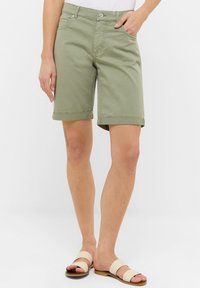 Shorts en coton vert clair avec ourlets roulés, poches avant et fermeture par bouton à l'avant, associés à des sandales beiges simples.