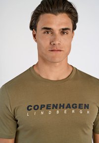Camiseta de algodón color verde oliva con mangas cortas, que presenta un texto audaz en blanco y negro "COPENHAGEN LINDBERGH" en el frente. Textura suave.