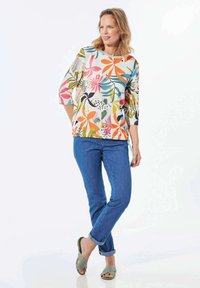 Mehrfarbige Blumenshirt mit Dreiviertelärmeln, kombiniert mit blauen Jeans und strukturierten grauen Sandalen. Leuchtende Muster auf hellen Hintergrund.